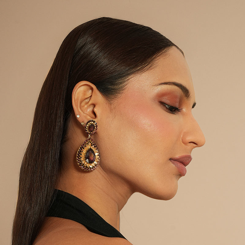 Woman wearing a pair of gold and amethyst colored earrings against a beige background